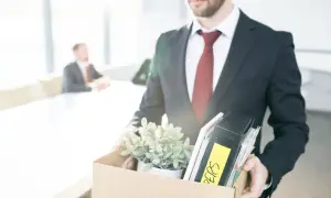 Un homme en costume quitte son bureau, portant une boîte remplie d'objets personnels et de dossiers. Il vient de déposer sa lettre de démission période d'essai.