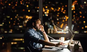 Un homme travaille tard, au téléphone pour une réclamation AXA, devant un paysage urbain nocturne illuminé.