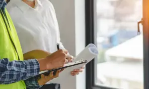 Un ouvrier en gilet jaune écrit sur un bloc-notes, tenant un rouleau de plans. Il s'agit d'une lettre de remboursement d'acompte pour des travaux non faits.