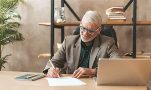 Homme âgé souriant écrivant sur un papier à son bureau, avec un ordinateur portable et une plante.