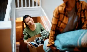 Un couple déménage, portant des cartons et une couverture dans un escalier. La femme, souriante, tient une boîte remplie d'objets. C'est une scène de déménagement dans un nouveau lieu.