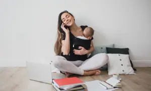 Femme au téléphone avec bébé en porte-bébé, entourée de livres et d'un ordinateur. Elle prépare sa lettre de prolongation de congé parental.