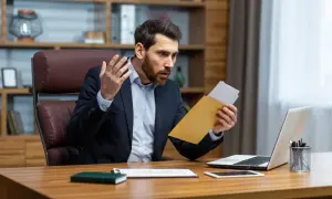 Homme surpris ouvrant une lettre de contestation de facture, assis à son bureau avec un ordinateur portable.