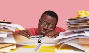 Un homme fatigué, entouré de piles de documents, semble avoir un avis de départ. Il repose sa tête sur un livre ouvert, l'air dépassé par la paperasse.