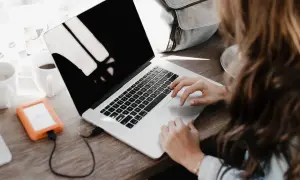 Une personne rédige une lettre de motivation française sur son ordinateur portable, avec une tasse de café et un disque dur externe à proximité.