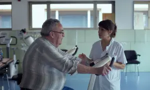 Une aide-soignante encourage un homme âgé sur un vélo d'appartement. Une scène qui pourrait figurer dans une lettre de motivation pour travailler auprès des personnes âgées.