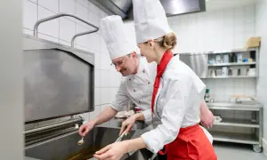 Deux chefs en cuisine, un homme et une femme, travaillent ensemble. L'homme, lunettes, semble superviser la jeune femme, qui porte un tablier rouge. Une scène parfaite pour une lettre de motivation second de cuisine.