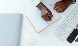 Un homme écrit une lettre réclamation frais bancaires dans un carnet. Un clavier et une souris sont à côté.