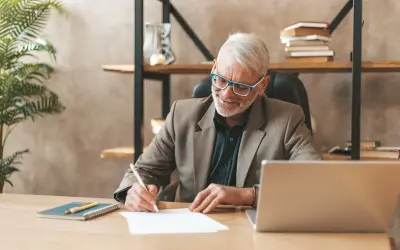 Homme âgé souriant écrivant sur un papier à son bureau, avec un ordinateur portable et une plante.