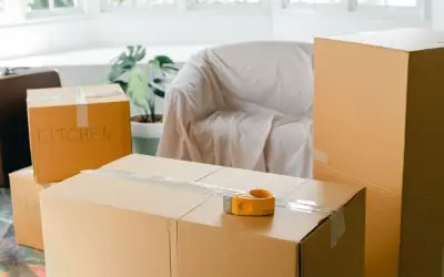 Cartons de déménagement prêts, dont un marqué "KITCHEN". Une valise et un fauteuil bâché complètent la scène.