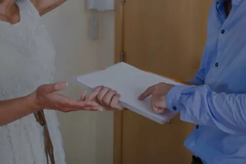 Une femme en robe blanche reçoit des documents d'un homme en chemise bleue.
