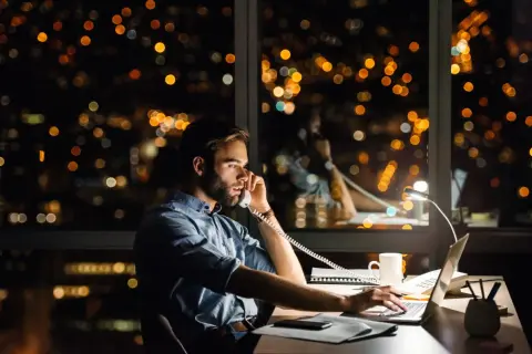 Un homme travaille tard, au téléphone pour une réclamation AXA, devant un paysage urbain nocturne illuminé.