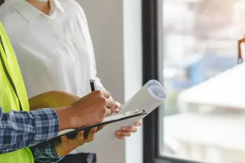 Un ouvrier en gilet jaune écrit sur un bloc-notes, tenant un rouleau de plans. Il s'agit d'une lettre de remboursement d'acompte pour des travaux non faits.