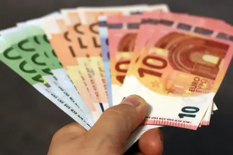 Main d'une personne tenant plusieurs billets en euros, dont des 10€ et 100€. Une scène évoquant une reclamation caisse epargne.