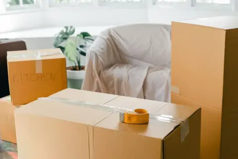 Cartons de déménagement prêts, dont un marqué "KITCHEN". Une valise et un fauteuil bâché complètent la scène.
