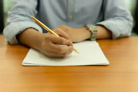 Main tenant une plume dorée, une personne rédige une lettre de motivation STAPS sur une feuille blanche.