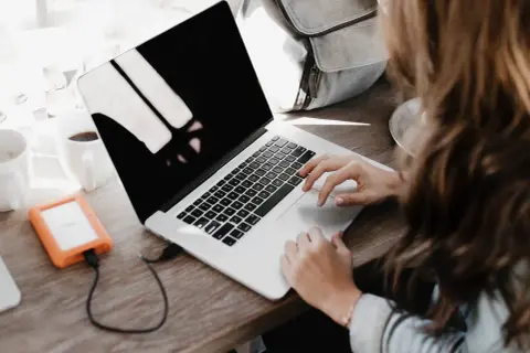 Une personne rédige une lettre de motivation française sur son ordinateur portable, avec une tasse de café et un disque dur externe à proximité.