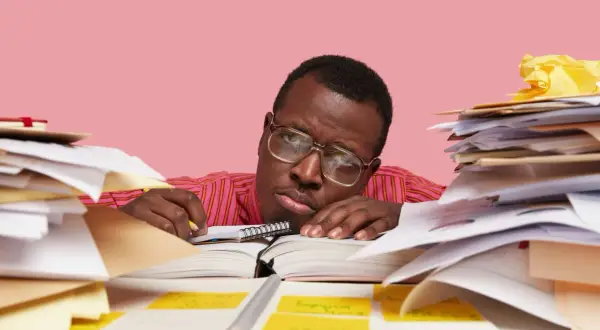 Un homme fatigué, entouré de piles de documents, semble dépassé par sa tâche. Il est peut-être en train de préparer un préavis foncia.