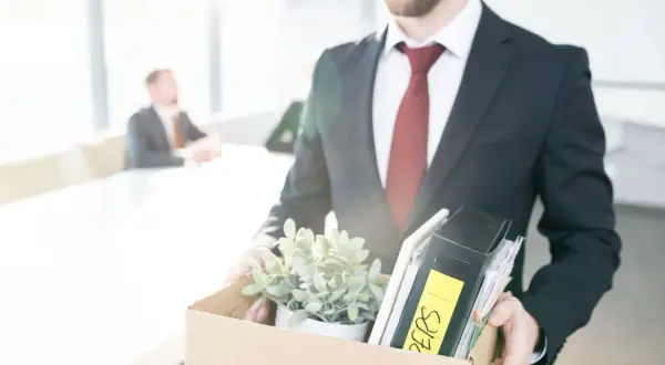 Un homme en costume quitte son bureau, portant une boîte remplie d'objets personnels et de dossiers. Il vient de déposer sa lettre de démission période d'essai.