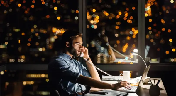 Un homme travaille tard, au téléphone pour une réclamation AXA, devant un paysage urbain nocturne illuminé.