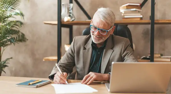 Homme âgé souriant écrivant sur un papier à son bureau, avec un ordinateur portable et une plante.
