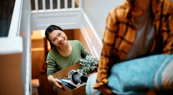 Un couple déménage, portant des cartons et une couverture dans un escalier. La femme, souriante, tient une boîte remplie d'objets. C'est une scène de déménagement dans un nouveau lieu.
