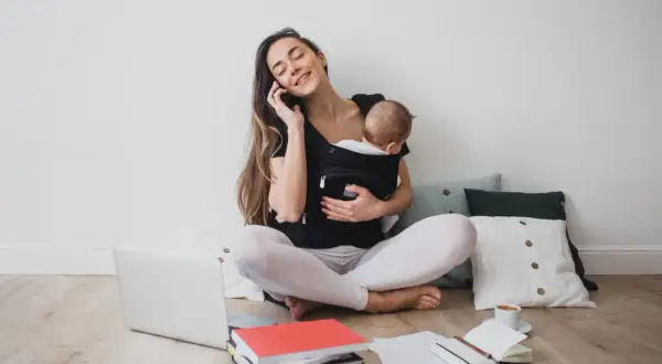 Femme au téléphone avec bébé en porte-bébé, entourée de livres et d'un ordinateur. Elle prépare sa lettre de prolongation de congé parental.