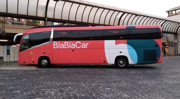 Un bus BlaBlaCar rouge et gris est garé sur une place pavée sous un auvent.