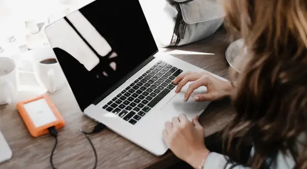 Une personne rédige une lettre de motivation française sur son ordinateur portable, avec une tasse de café et un disque dur externe à proximité.