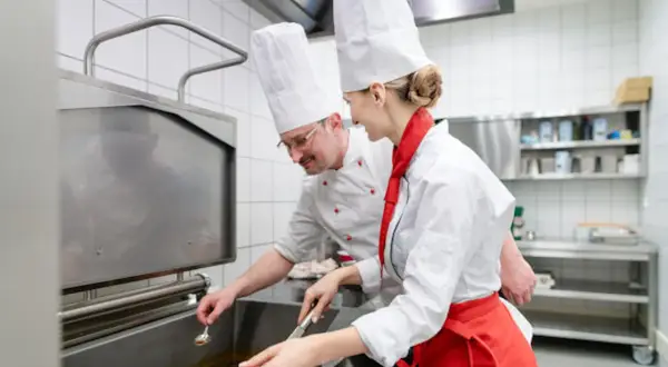 Deux chefs en cuisine, un homme et une femme, travaillent ensemble. L'homme, lunettes, semble superviser la jeune femme, qui porte un tablier rouge. Une scène parfaite pour une lettre de motivation second de cuisine.