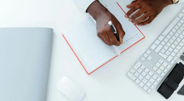 Un homme écrit une lettre réclamation frais bancaires dans un carnet. Un clavier et une souris sont à côté.