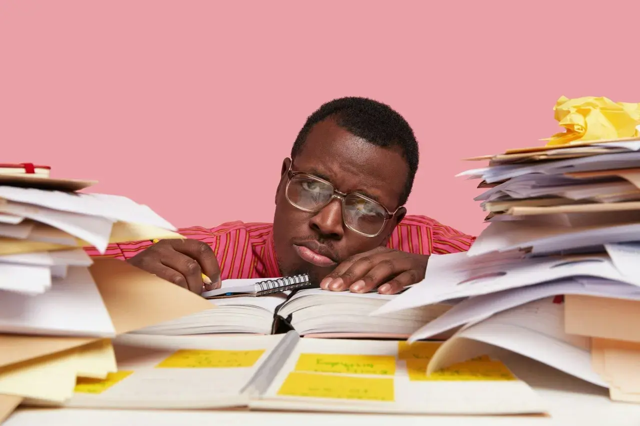 Un homme fatigué, entouré de piles de documents, semble dépassé par sa tâche. Il est peut-être en train de préparer un préavis foncia.