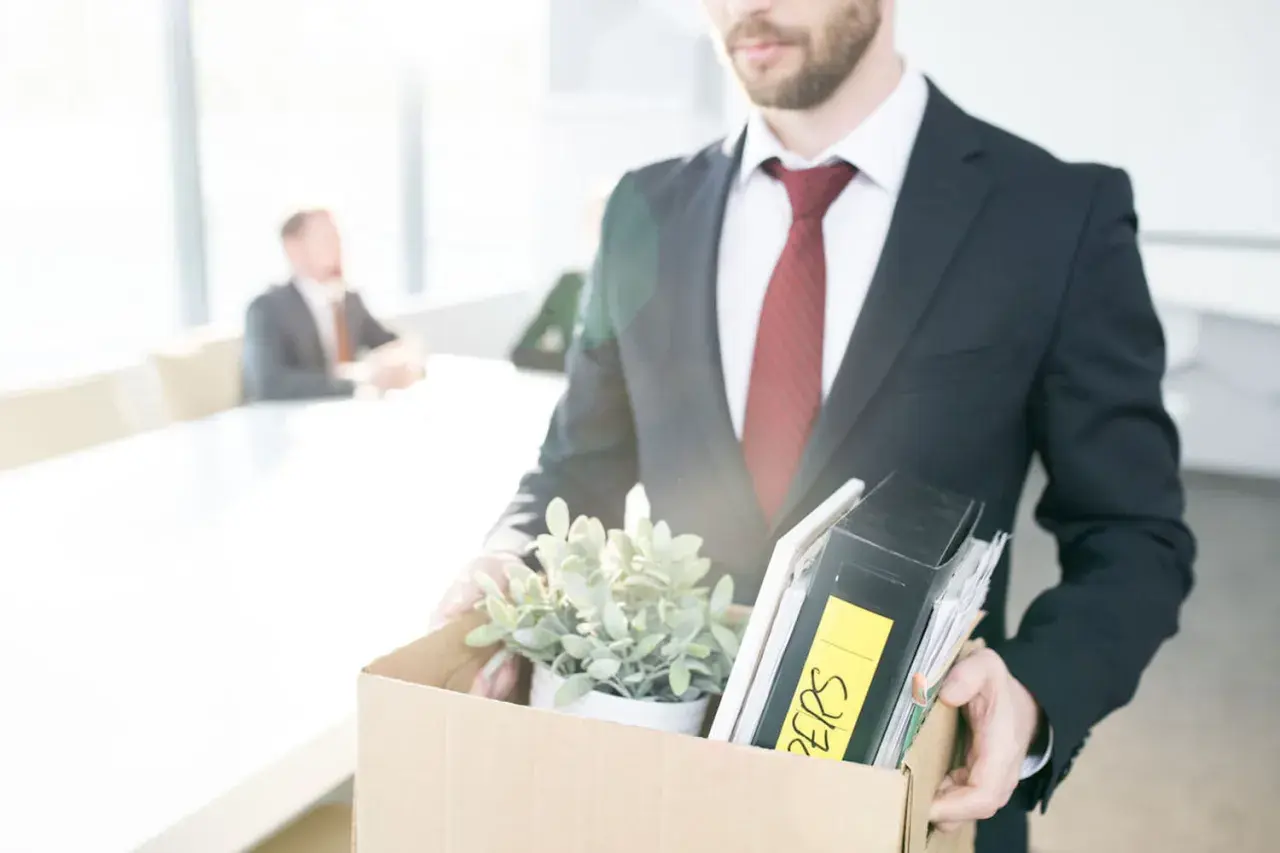 Un homme en costume quitte son bureau, portant une boîte remplie d'objets personnels et de dossiers. Il vient de déposer sa lettre de démission période d'essai.