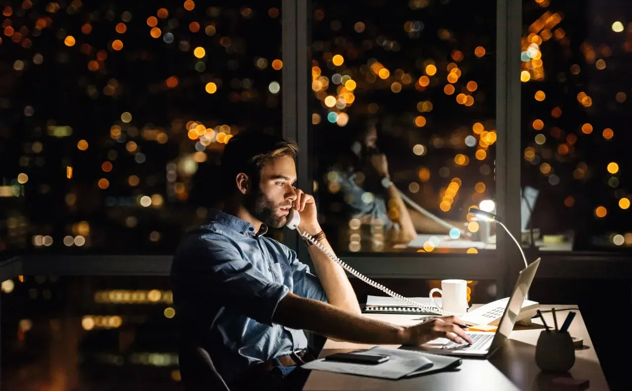 Un homme travaille tard, au téléphone pour une réclamation AXA, devant un paysage urbain nocturne illuminé.