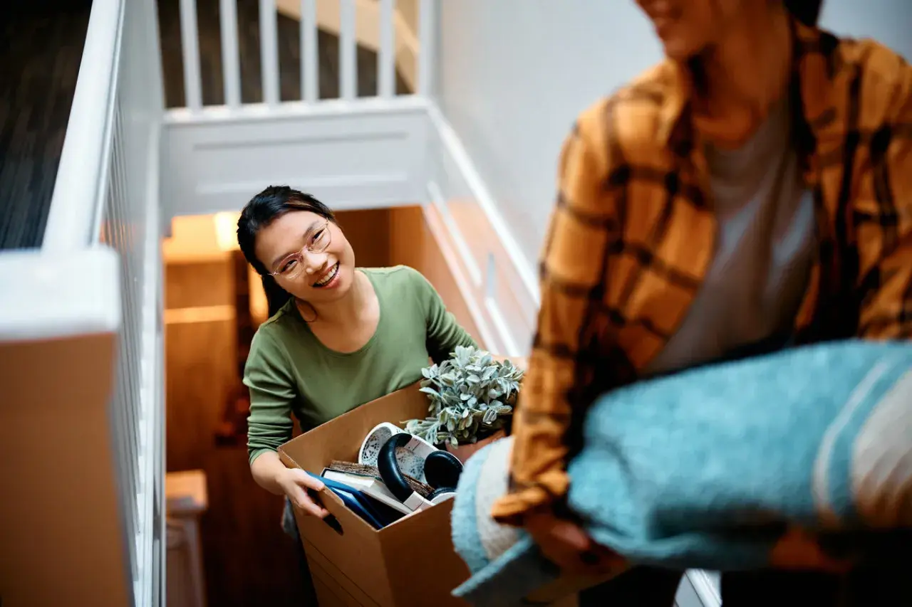 Un couple déménage, portant des cartons et une couverture dans un escalier. La femme, souriante, tient une boîte remplie d'objets. C'est une scène de déménagement dans un nouveau lieu.