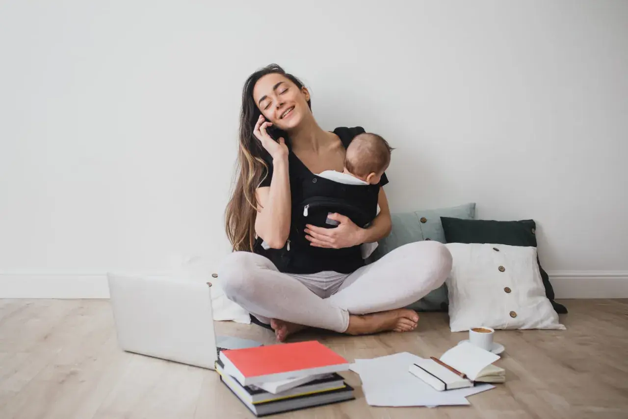 Femme au téléphone avec bébé en porte-bébé, entourée de livres et d'un ordinateur. Elle prépare sa lettre de prolongation de congé parental.