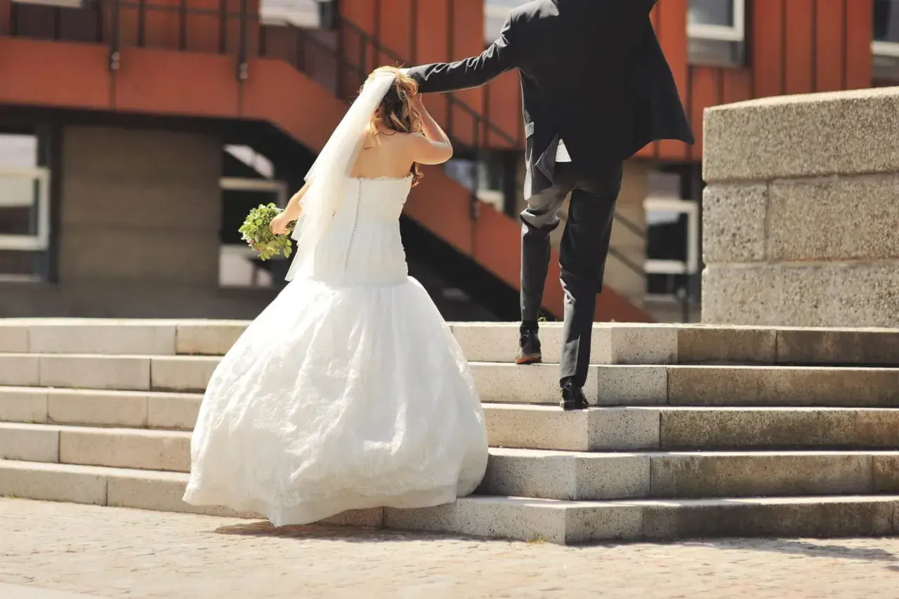 Un couple monte les marches, la mariée en robe blanche tenant un bouquet, le marié en costume noir. Une scène symbolisant leur union, peut-être après une lettre mariage mairie.