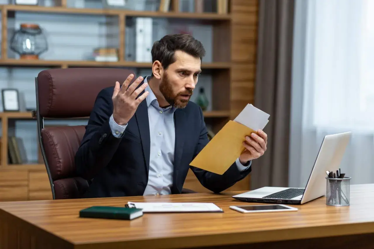 Homme surpris ouvrant une lettre de contestation de facture, assis à son bureau avec un ordinateur portable.