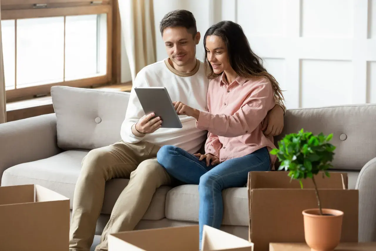 Couple sur un canapé, regardant une tablette. Des cartons de déménagement sont autour d'eux, peut-être pour une lettre pour quitter son logement sans préavis.