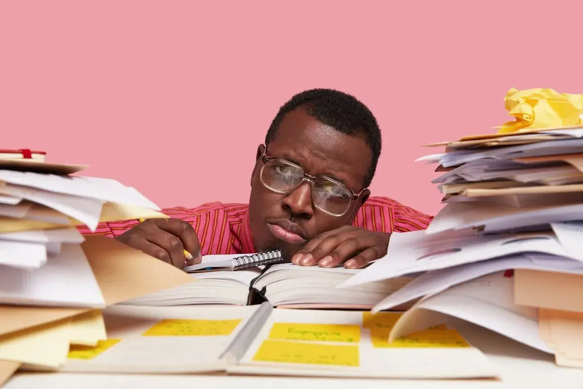 Un homme fatigué, entouré de piles de documents, semble avoir un avis de départ. Il repose sa tête sur un livre ouvert, l'air dépassé par la paperasse.