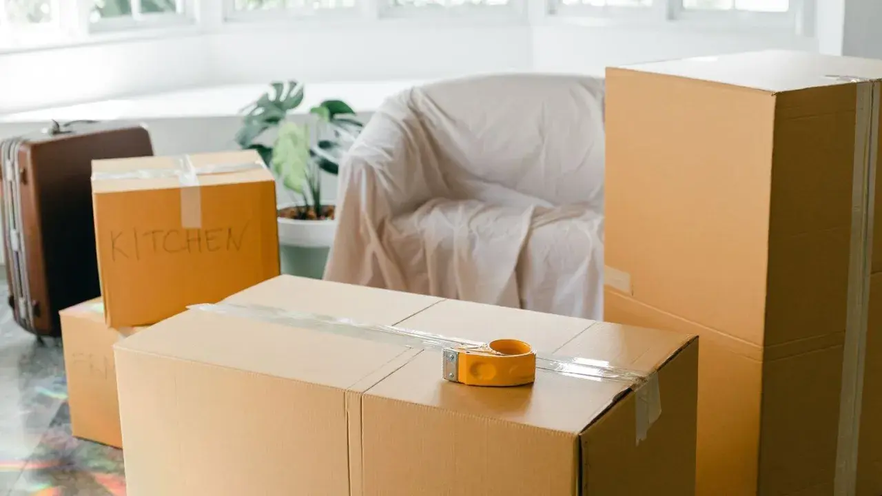 Cartons de déménagement prêts, dont un marqué "KITCHEN". Une valise et un fauteuil bâché complètent la scène.