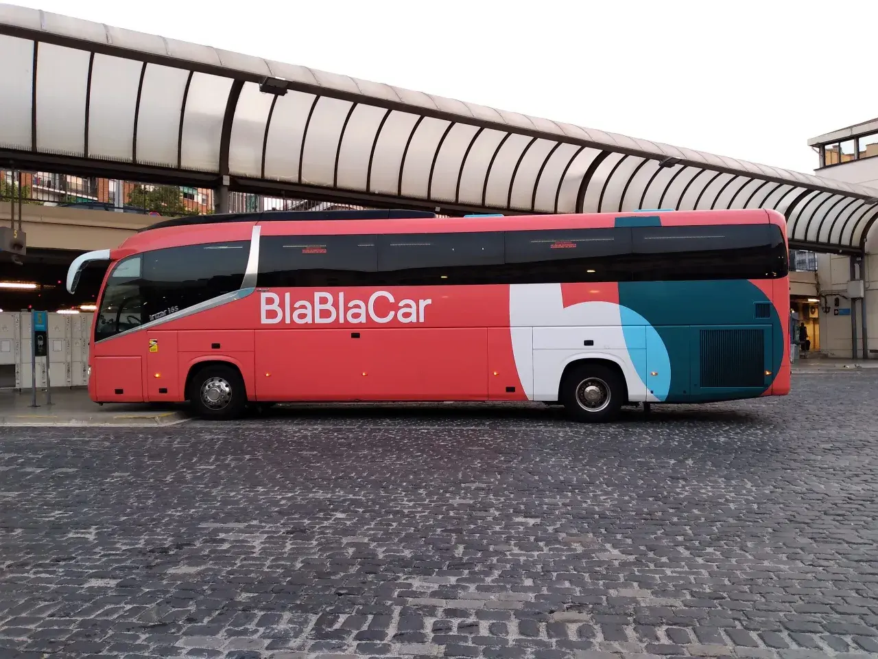 Un bus BlaBlaCar rouge et gris est garé sur une place pavée sous un auvent.