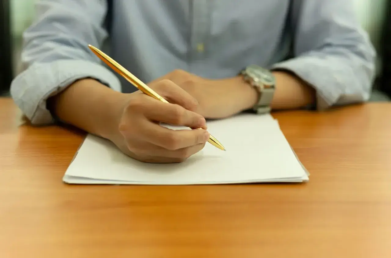 Main tenant une plume dorée, une personne rédige une lettre de motivation STAPS sur une feuille blanche.