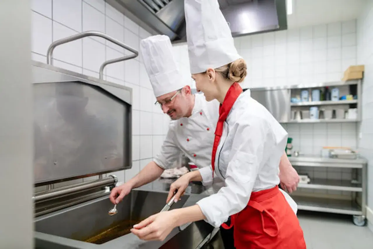 Deux chefs en cuisine, un homme et une femme, travaillent ensemble. L'homme, lunettes, semble superviser la jeune femme, qui porte un tablier rouge. Une scène parfaite pour une lettre de motivation second de cuisine.