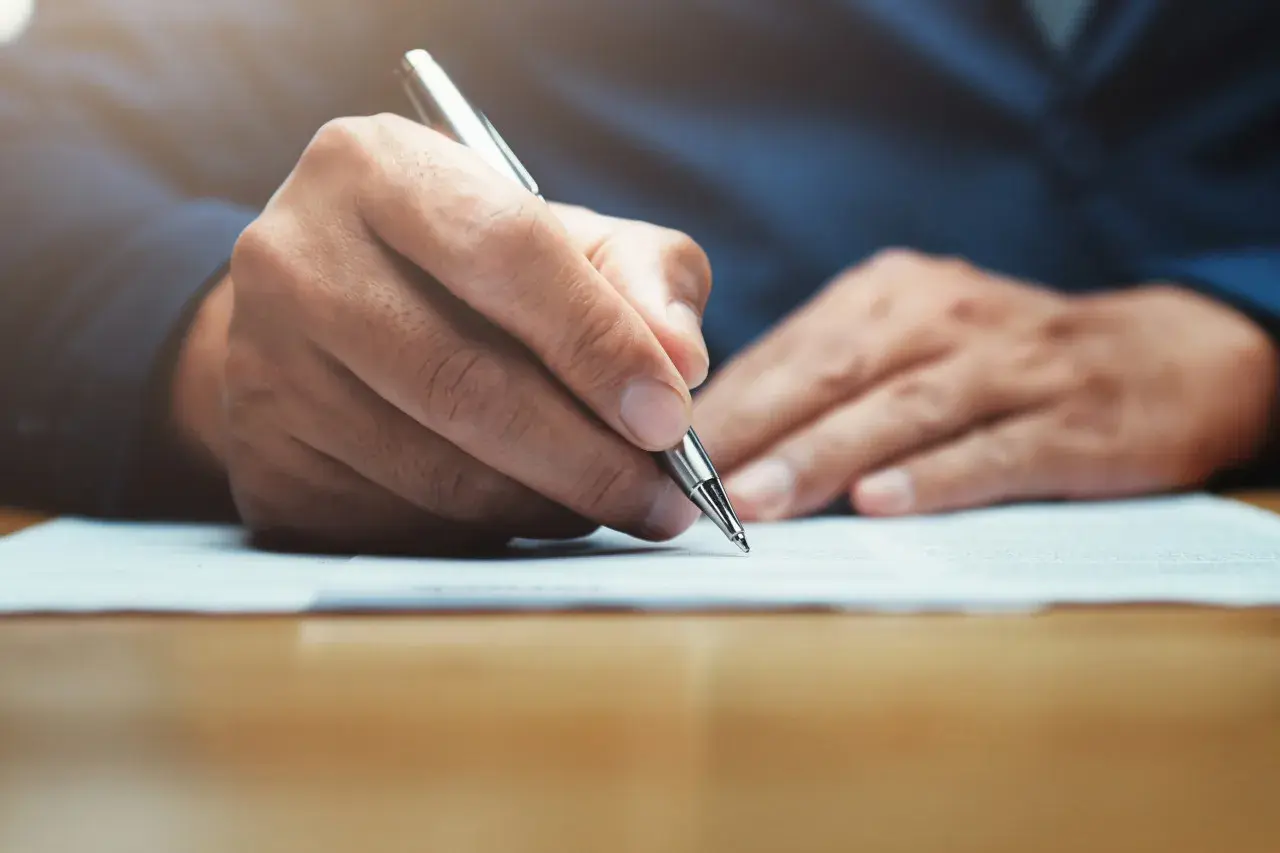 Main tenant rédigeant une lettre, peut-être un modèle lettre CARSAT, avec un stylo argenté sur un document.