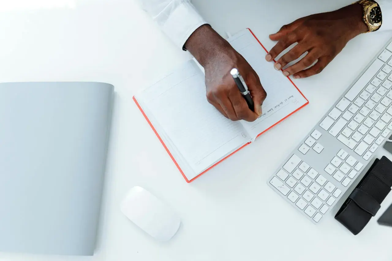 Un homme écrit une lettre réclamation frais bancaires dans un carnet. Un clavier et une souris sont à côté.