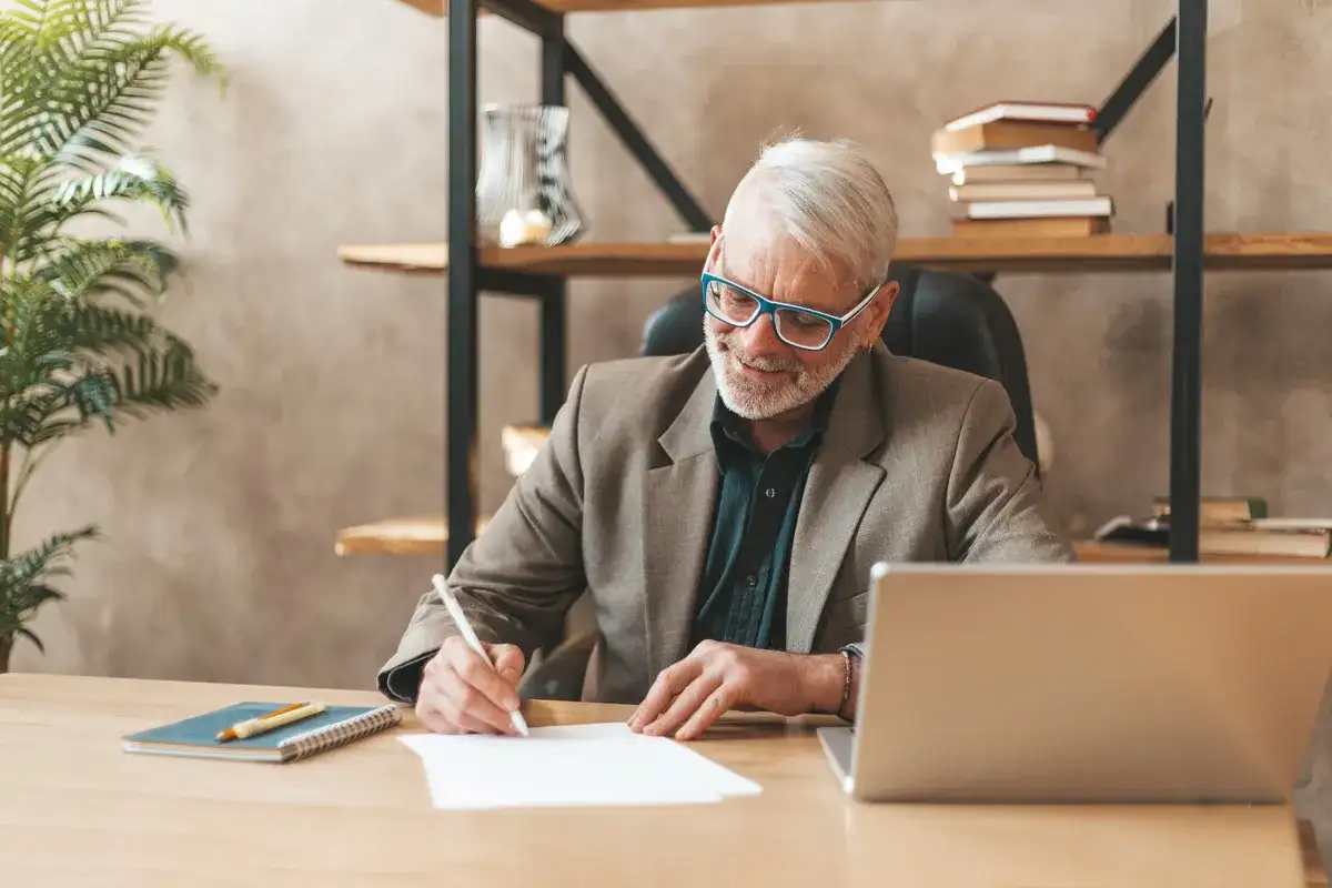Homme âgé souriant écrivant sur un papier à son bureau, avec un ordinateur portable et une plante.