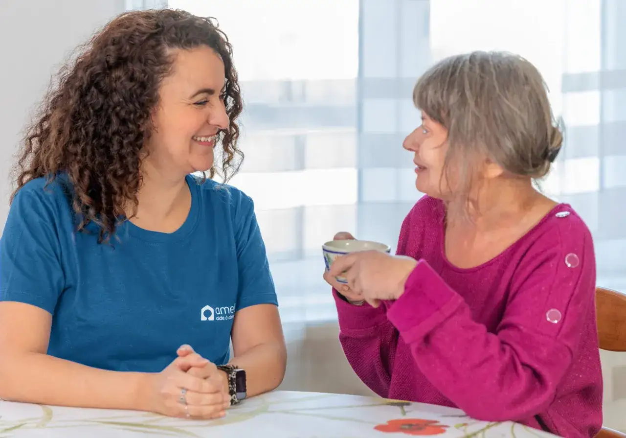 Une aide à domicile souriante discute avec une personne âgée autour d'une tasse. Une belle image pour une lettre de motivation pour travailler auprès des personnes âgées.