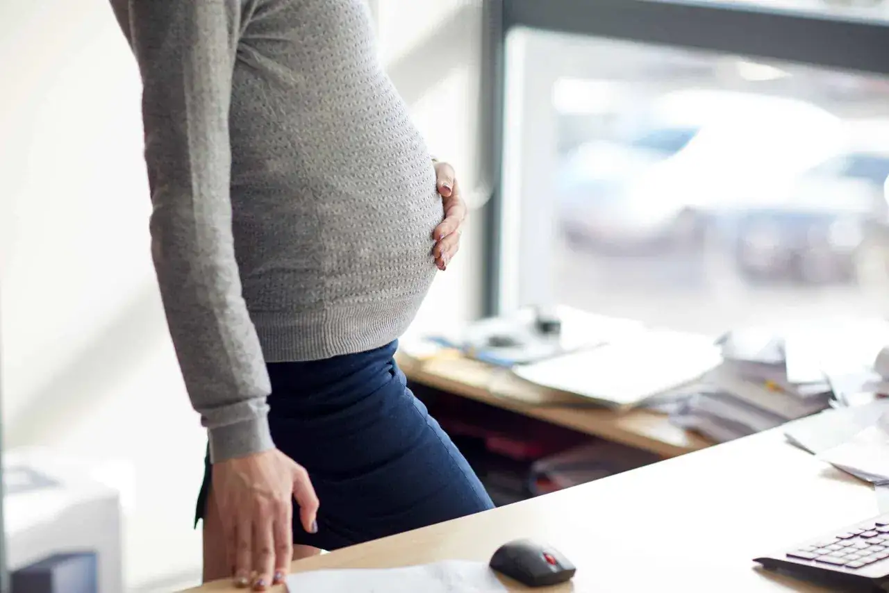 Femme enceinte tenant son ventre, pr&egrave;s d'un bureau encombr&eacute;. Elle pourrait pr&eacute;parer une lettre de d&eacute;mission pendant cong&eacute; maternit&eacute;.