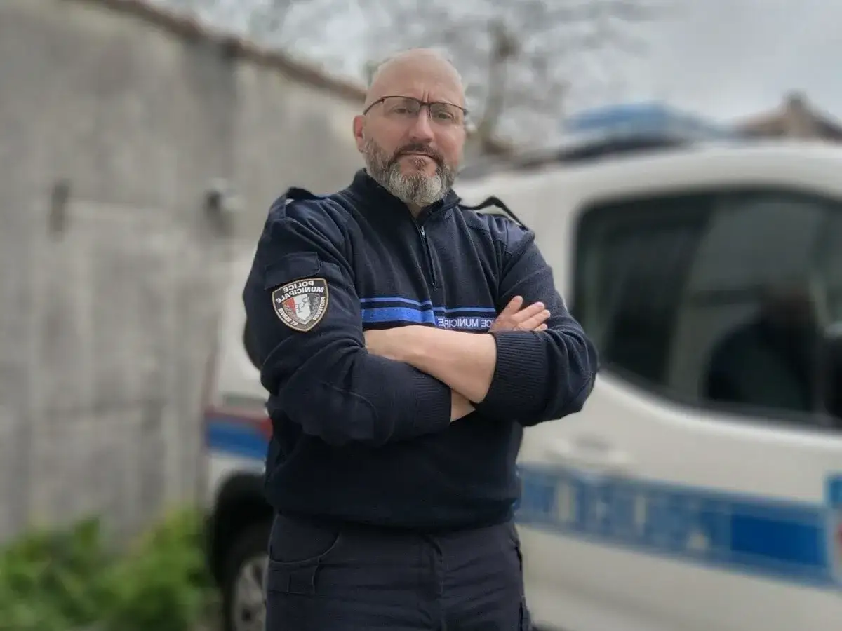 Policier municipal, bras crois&eacute;s, pr&ecirc;t pour une lettre de motivation police municipale mutation. V&eacute;hicule de service flout&eacute; en arri&egrave;re-plan.